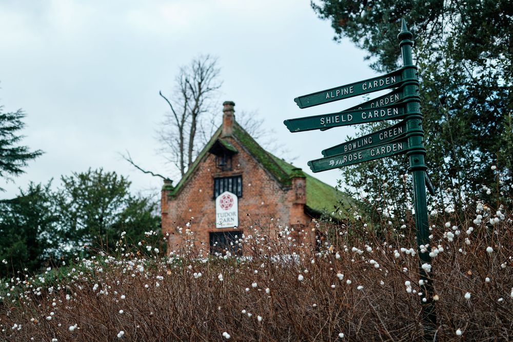 Brick garden building with moss-covered roof beside a green directional signpost in a lush botanical garden setting.