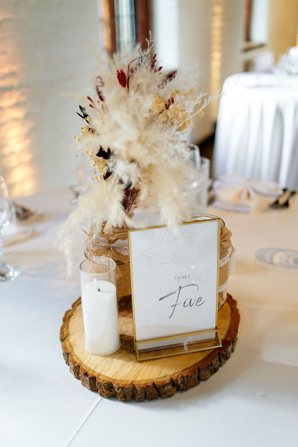 Rustic wedding table centerpiece with pampas grass, dried flowers, a white candle, and a gold-framed Table Five sign on a wood slice.