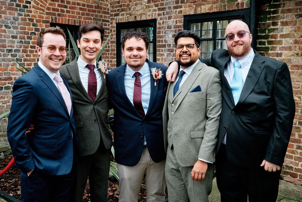 Five men in suits smiling together outdoors near a brick wall at a formal event or wedding.
