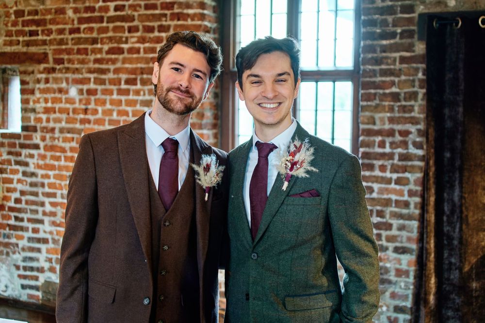 Two grooms in matching brown tweed suits with burgundy ties and boutonnieres pose together at a wedding venue.