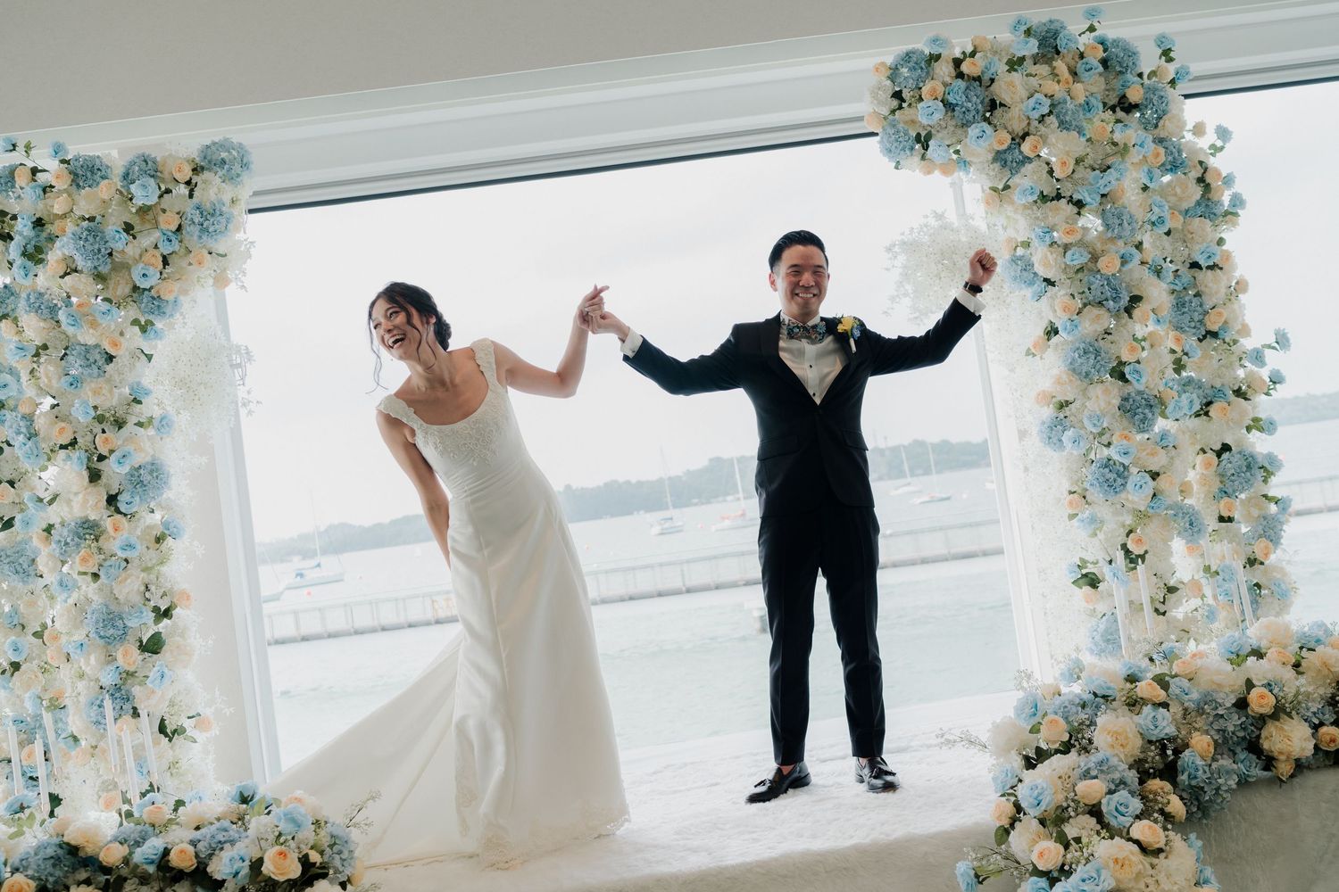 Joyful bride and groom celebrating under a floral arch with blue and white flowers, overlooking a scenic waterfront view.
