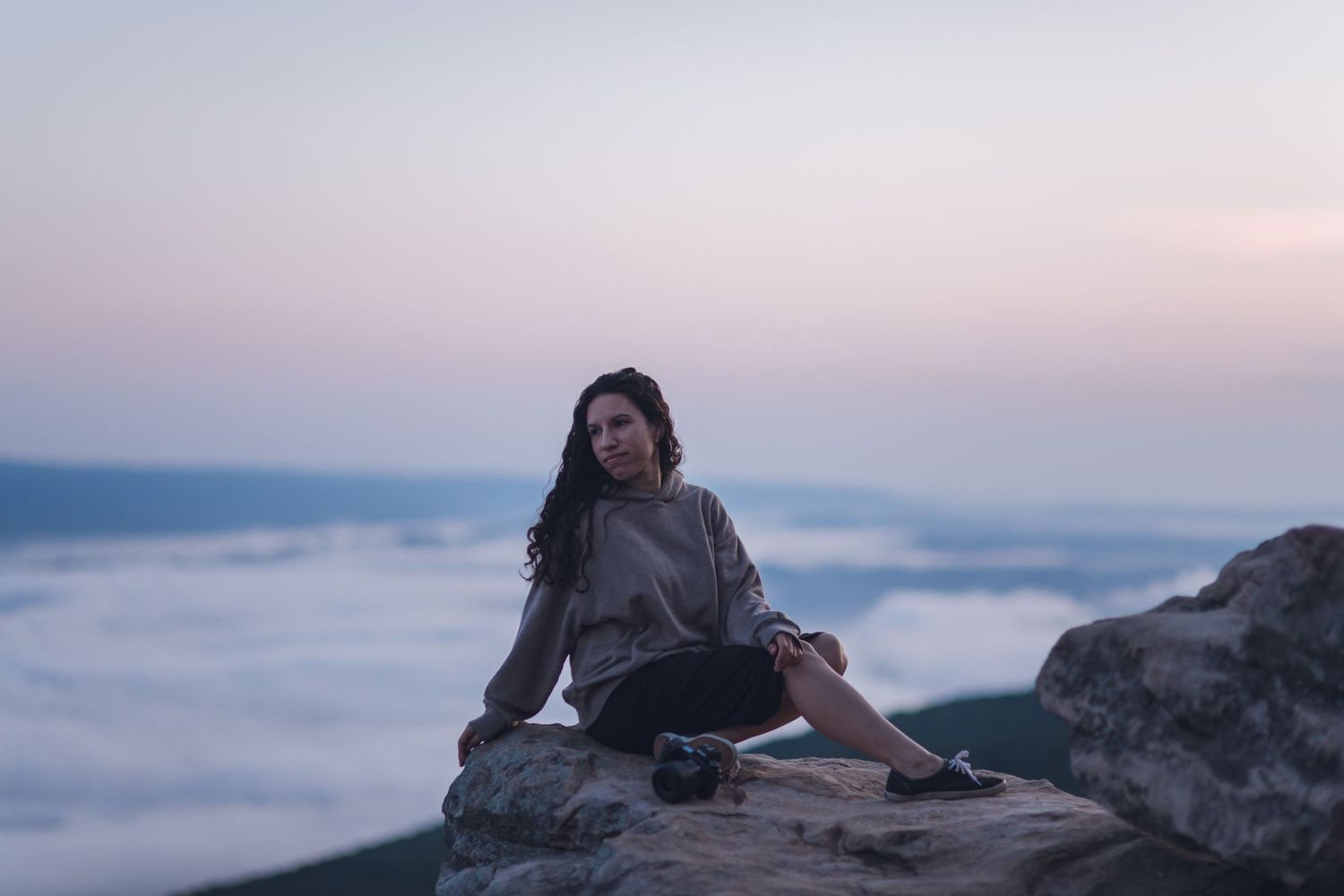 A person sits on a rocky mountain summit above a sea of clouds during sunset in a tranquil scene.