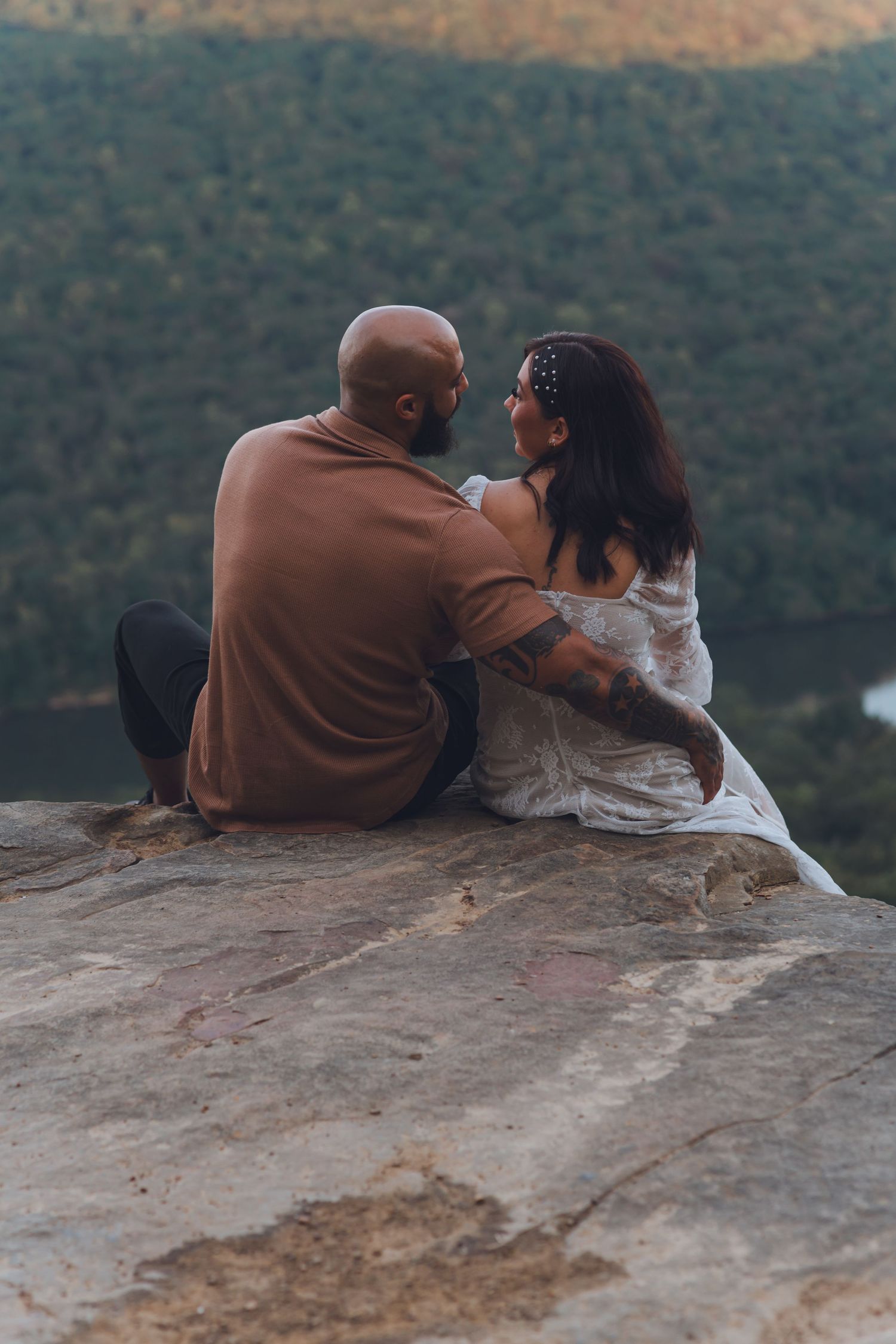 A couple sits together on a rocky cliff edge overlooking mountainous terrain during golden hour.