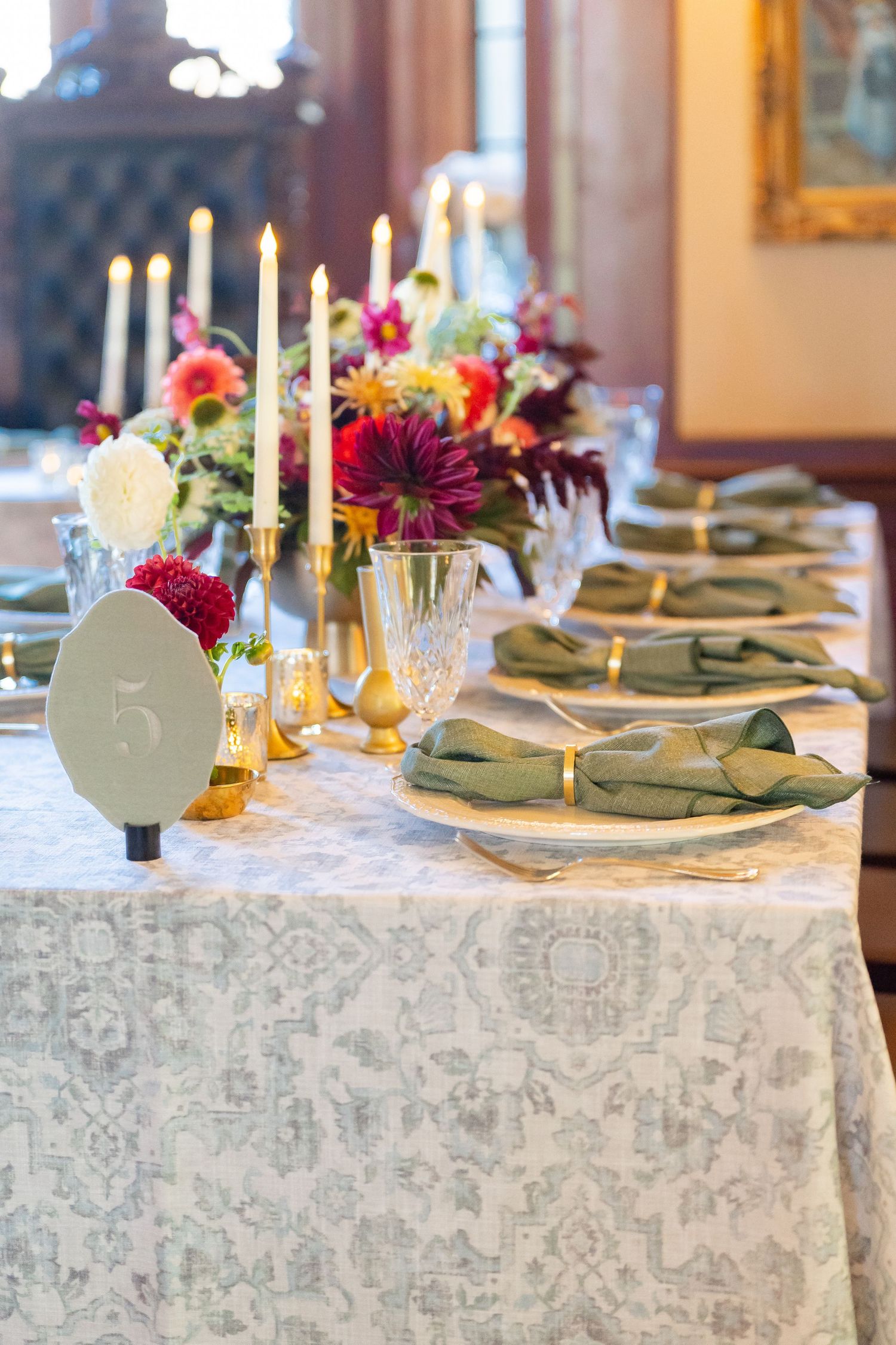 Elegant wedding table setting with green napkins, gold rings, crystal glasses, candles, and colorful floral centerpieces on patterned linen.