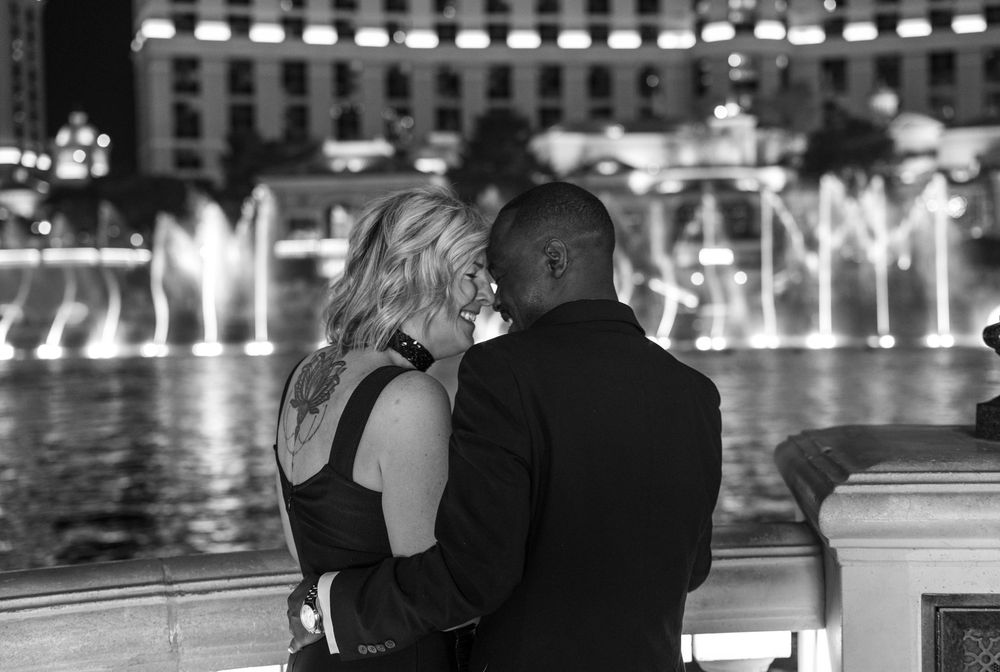 Couple embracing romantically in front of illuminated fountains at night in black and white photo.