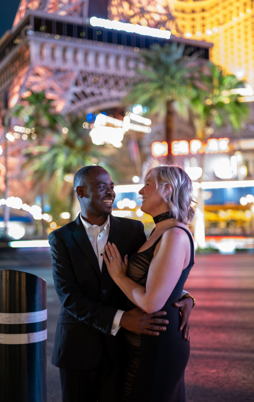Couple embracing and smiling on the Las Vegas Strip at night with the Eiffel Tower replica glowing behind them.