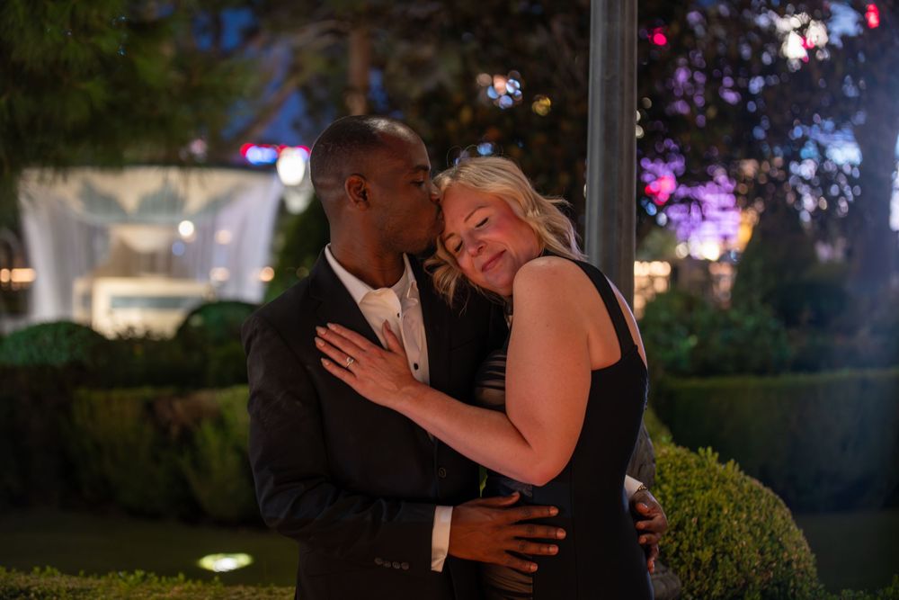 Couple embracing romantically outdoors at night with garden lights and greenery in background.