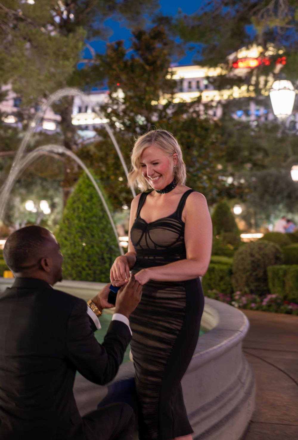 Man on one knee proposing to smiling woman near fountain in Las Vegas at night.