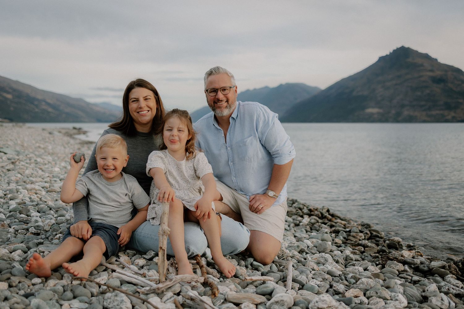 Family of four smiling on a rocky lakeshore with mountains in the background.