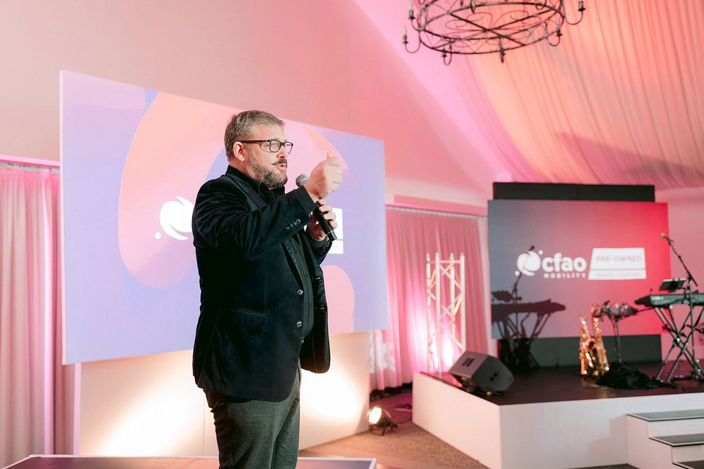 Man speaking at a pink-lit event stage with a microphone, presentation screen and Octao branding visible in background.
