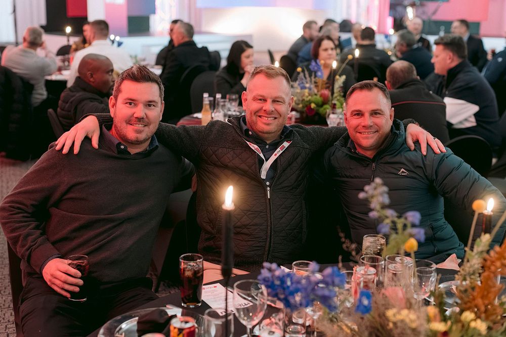 Three men smile together at a candlelit dinner event table with floral centerpieces and guests in background.