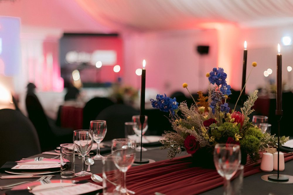 Elegant event table setting with black candles, colorful floral centerpiece, wine glasses, and pink lighting in a banquet hall.