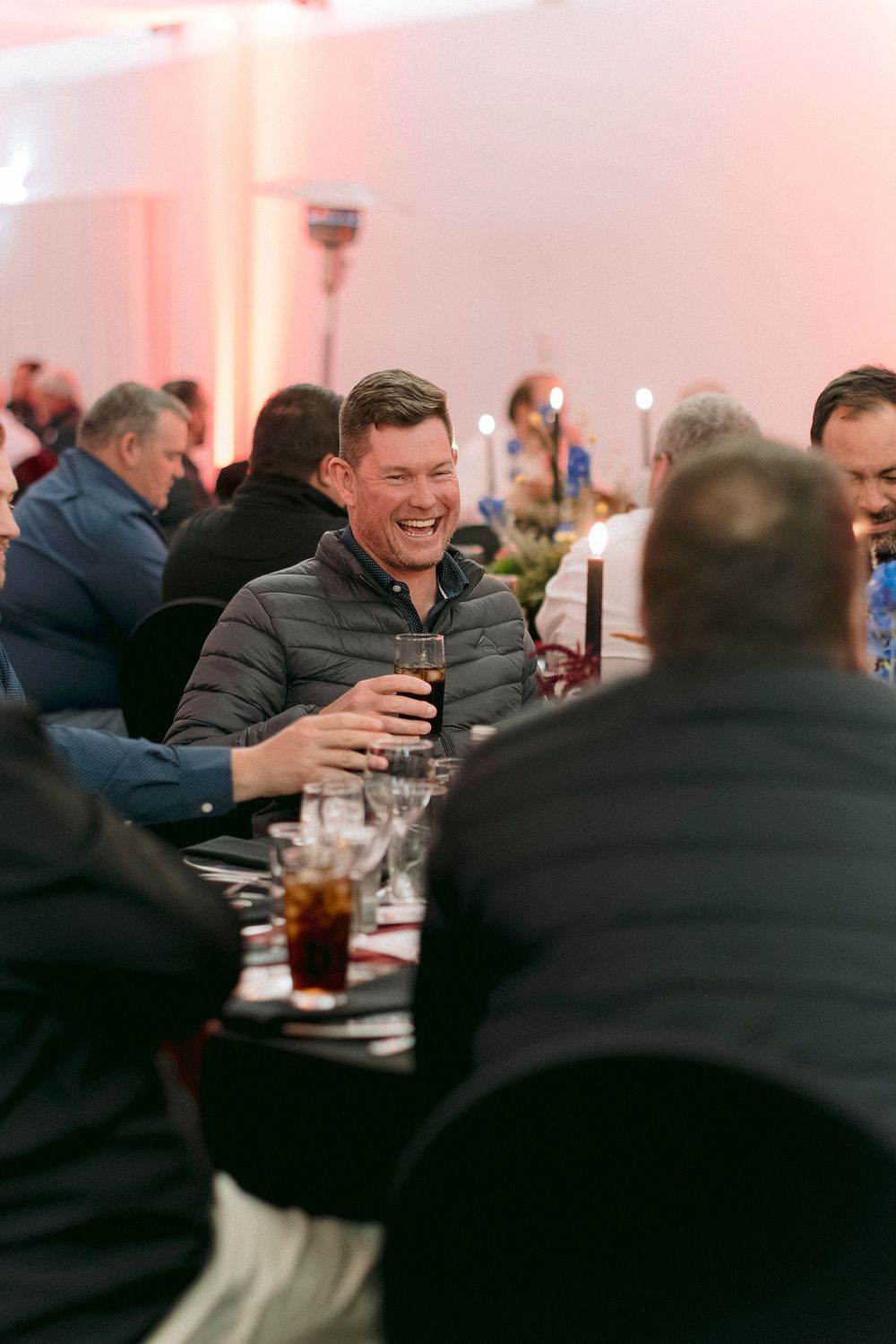 Man laughing and holding a drink at a formal dinner event with guests seated at tables.
