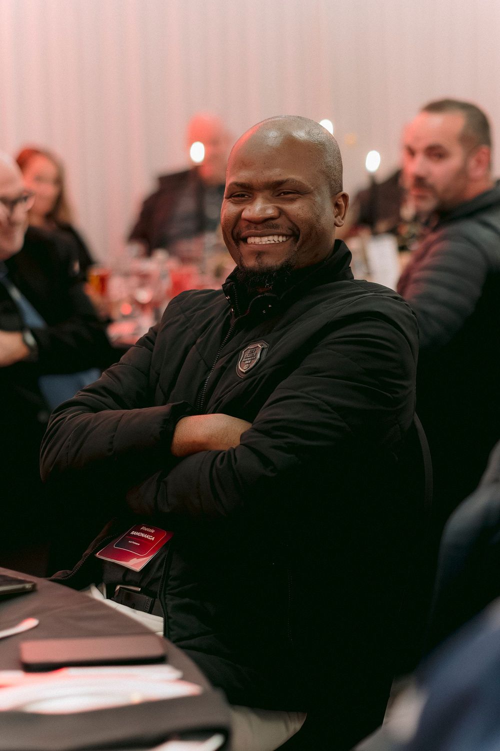 Smiling bald man in black jacket sits with arms crossed at an indoor event, with guests and red decor in background.