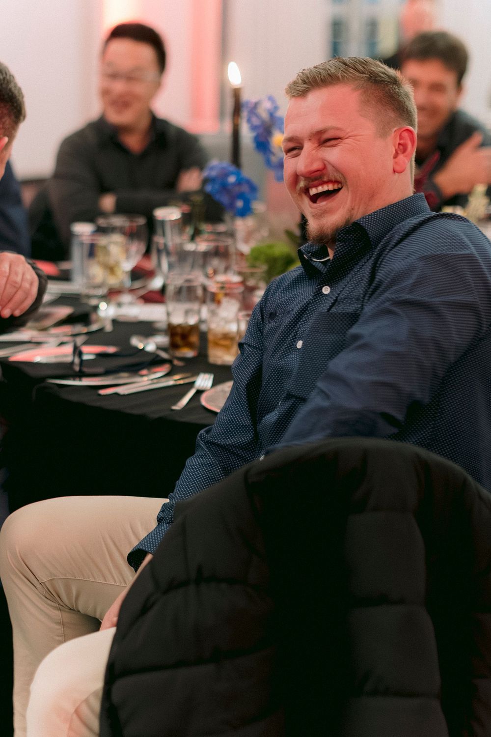 A man in a blue shirt laughing joyfully at a dinner event with guests seated at a decorated table.