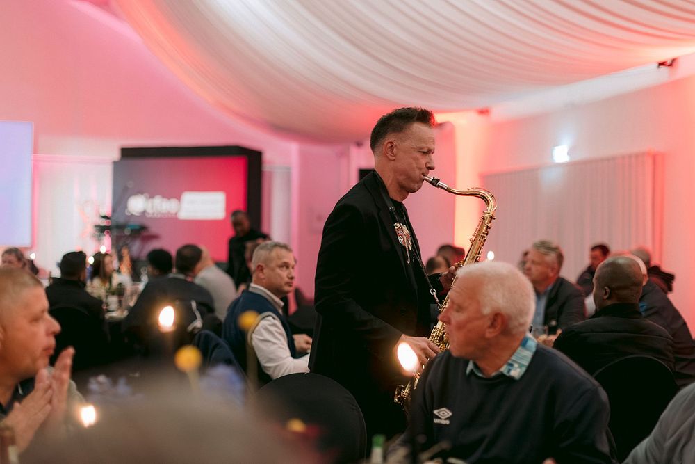 A saxophonist performs live at an elegant indoor event dinner with guests seated at candlelit tables under pink ambient lighting.