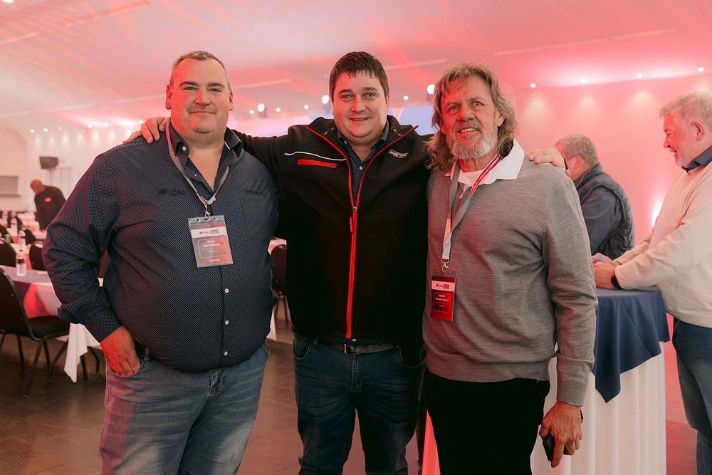 Three men pose together smiling at an indoor event with red ambient lighting and guests in the background.