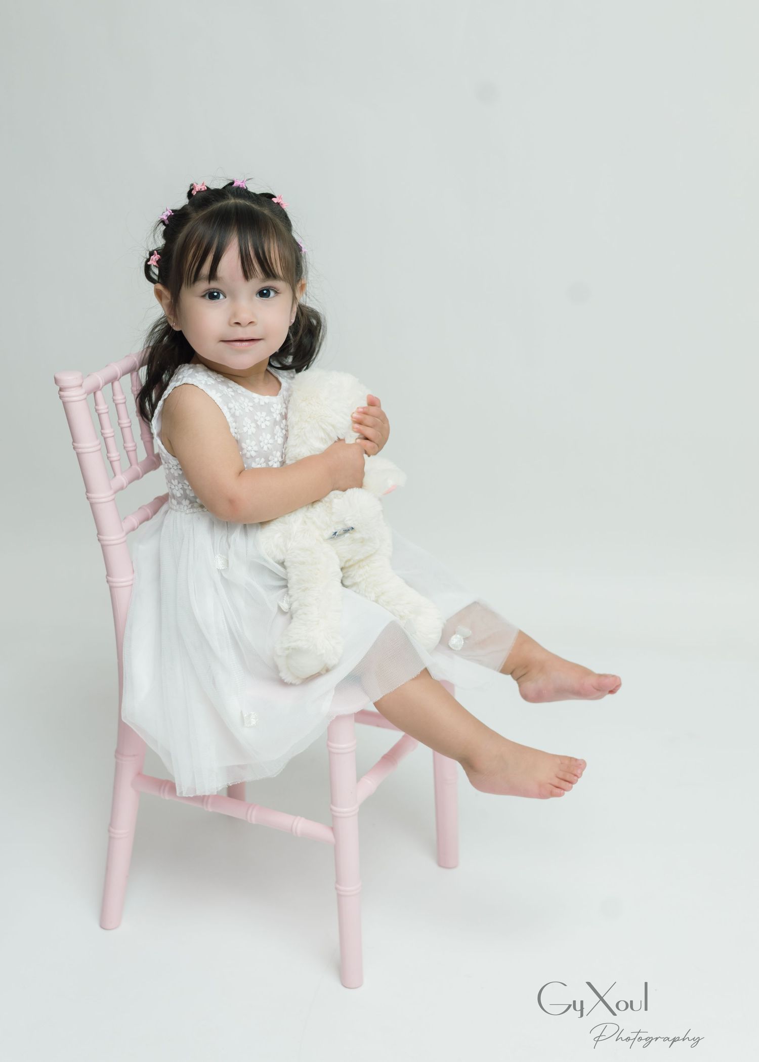 Young girl in white dress sits on pink chair hugging a white stuffed animal toy in a studio photo.