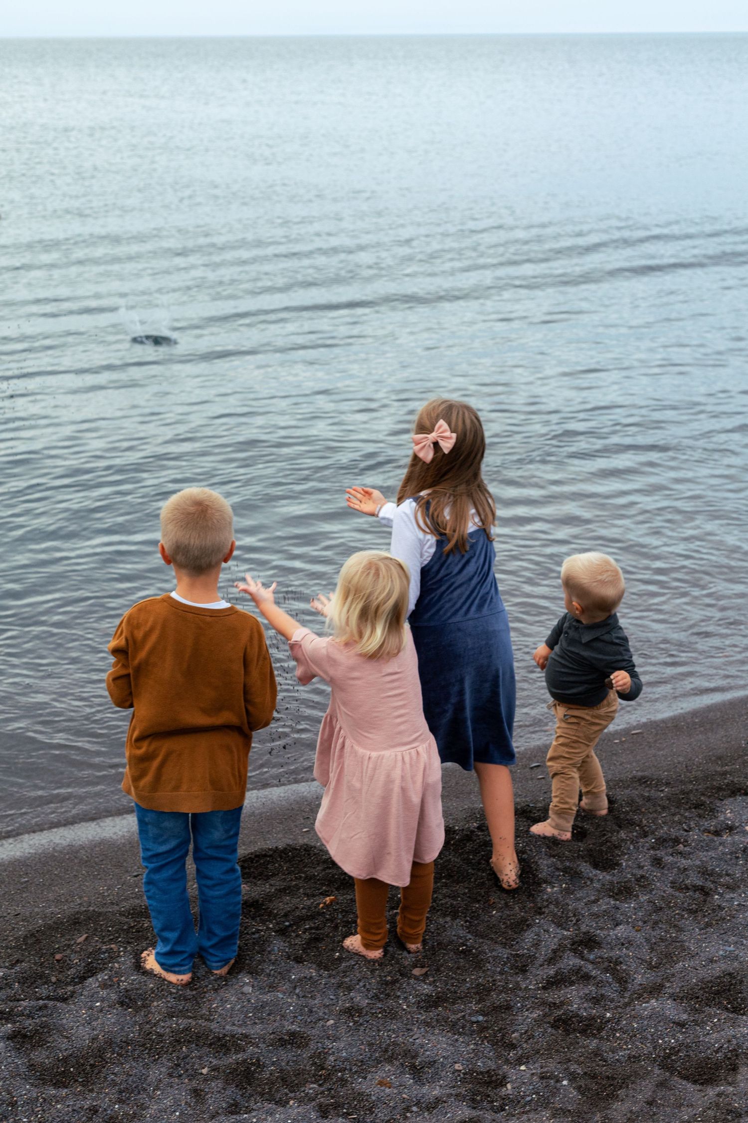 Four young children standing on a rocky beach shore, pointing at the calm ocean water on an overcast day.