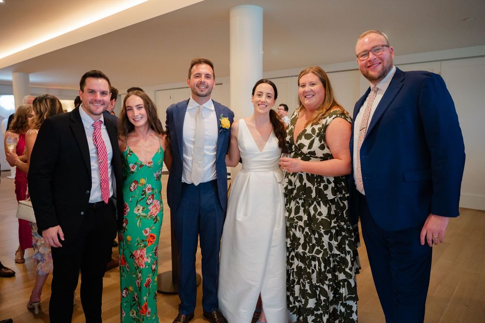 A group of wedding guests gather in a reception hall wearing formal attire including suits, dresses and evening wear.