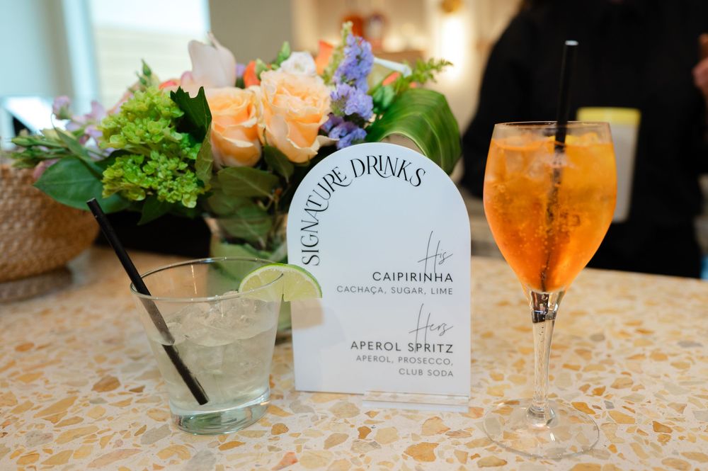 A series of tropical cocktails lined up on a lacy tablecloth with garnishes and a bar menu displaying drink options.