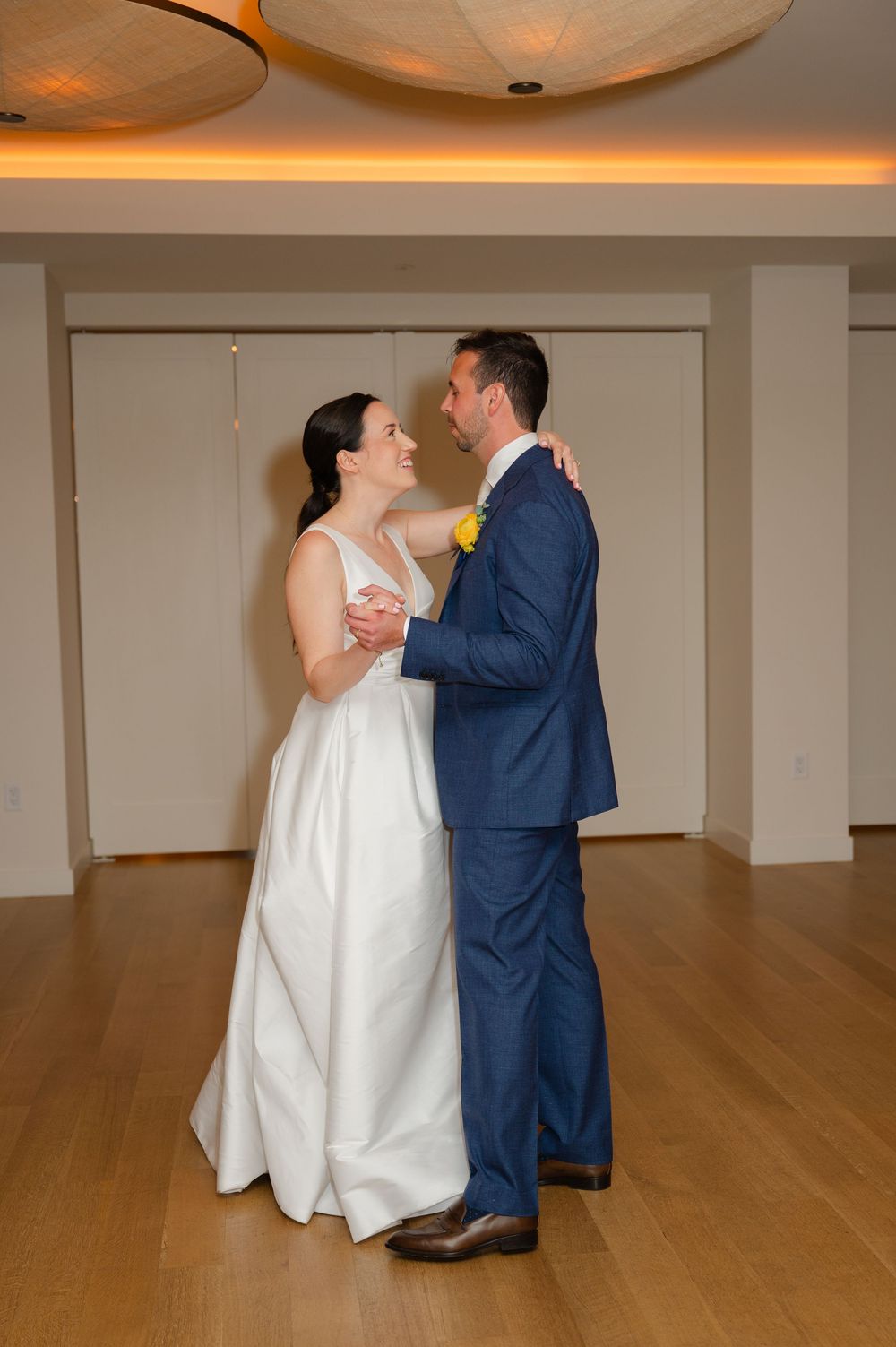 A newly married couple shares their first dance in an elegant room with hardwood floors and warm lighting.