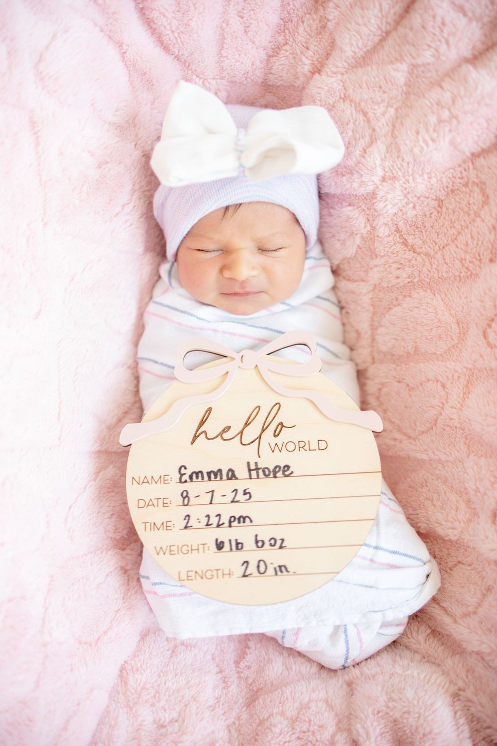 Newborn baby Emma Hope swaddled in striped wrap with bow hat holding hello world birth announcement sign.