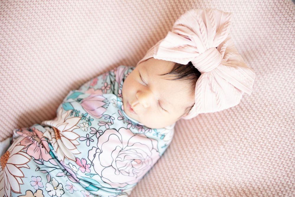 Newborn baby girl sleeping in colorful floral swaddle with large pink bow turban headband on pink blanket.