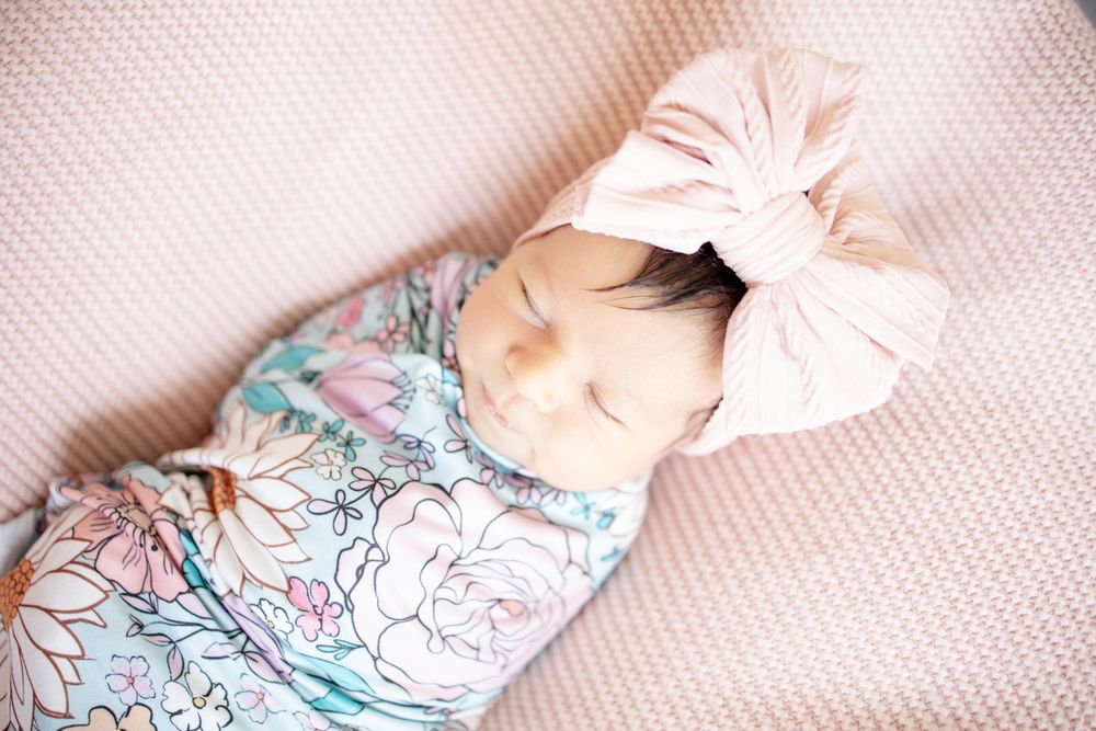 Sleeping newborn baby girl in floral swaddle with pink bow headband lying on textured pink blanket.