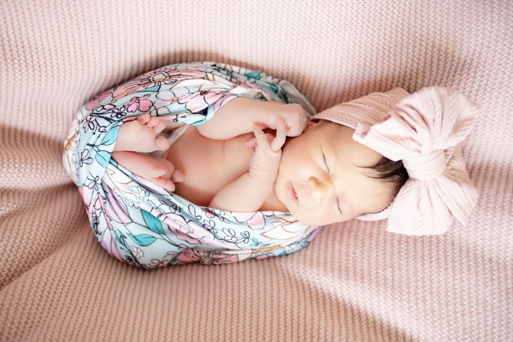 Newborn baby girl unswaddled in floral blanket with pink bow headband showing tiny hands on pink blanket.