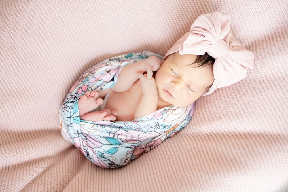 Newborn baby girl sleeping in floral swaddle with pink bow headband posed on textured pink blanket.