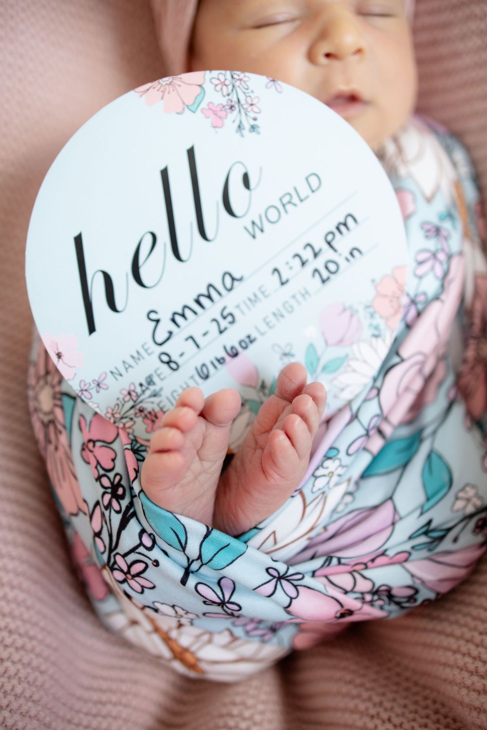 Newborn baby Emma holds hello world floral birth announcement sign showing tiny feet with stats on pink blanket.