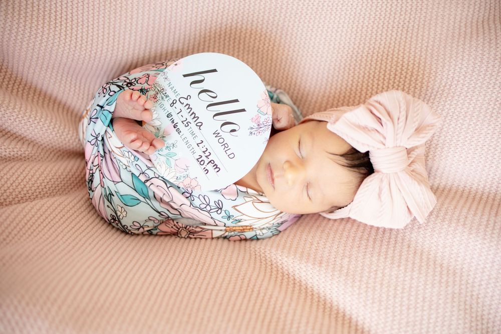 Sleeping newborn baby girl swaddled in floral wrap with pink bow headband and hello world birth announcement sign.