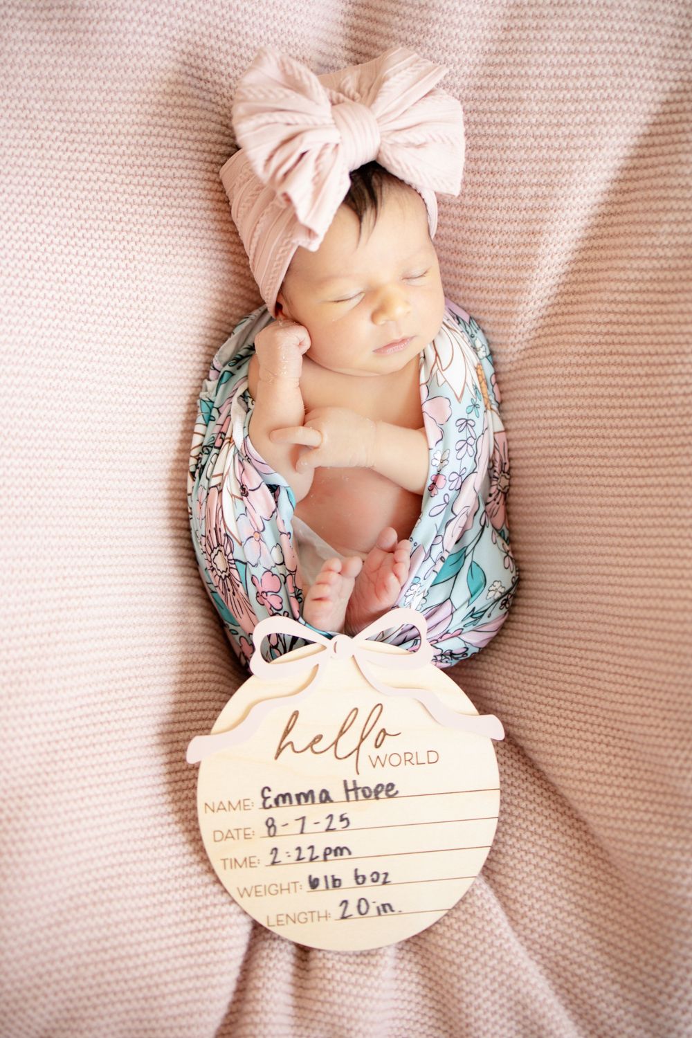 Newborn baby girl Emma Hope swaddled in floral wrap with pink bow headband beside hello world wooden birth stats sign.