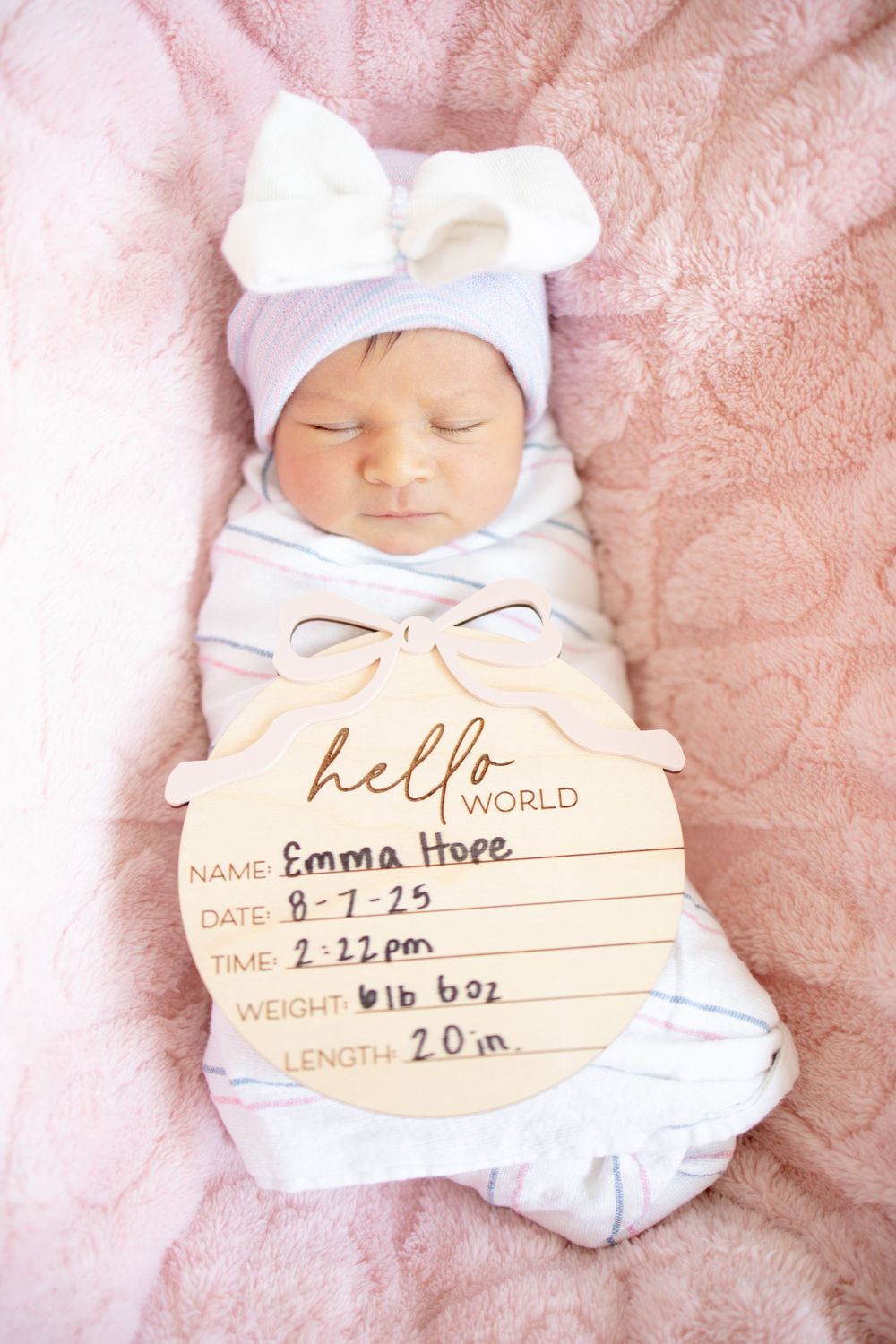 Newborn baby girl Emma swaddled in white with bow hat holds hello world birth announcement sign on pink blanket.