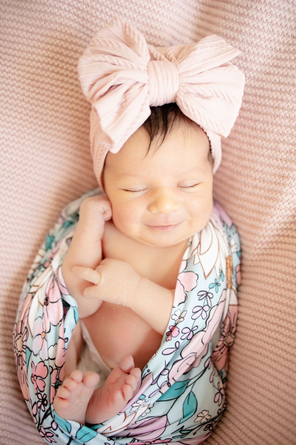 Smiling newborn baby girl swaddled in colorful floral wrap wearing a large pink bow headband on pink blanket.