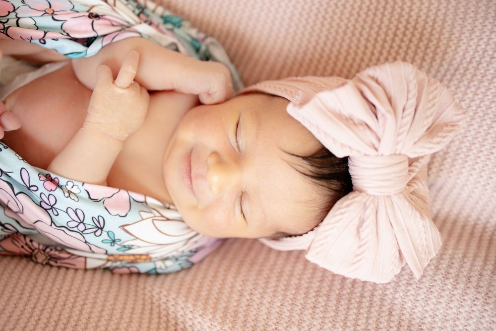 Close-up of smiling newborn baby girl in floral swaddle with pink bow headband, hands near face.