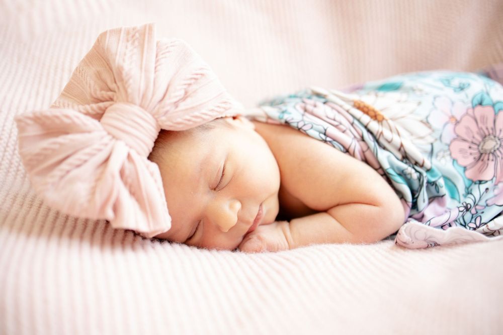 Peaceful sleeping newborn baby girl in floral swaddle with pink bow headband lying on soft pink bedding.
