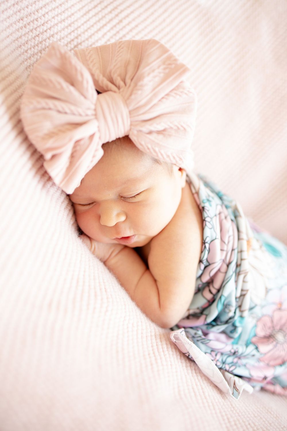 Newborn baby girl sleeping in floral swaddle with large pink ribbed bow headband on soft pink blanket.
