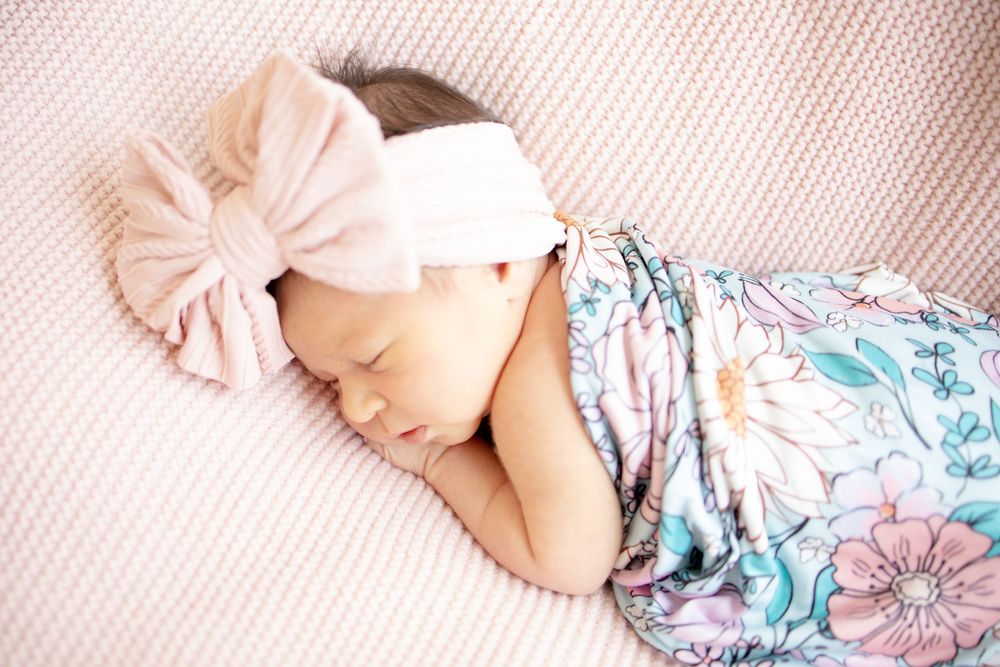 Sleeping newborn baby girl in colorful floral swaddle with large pink velvet bow headband on pink blanket.