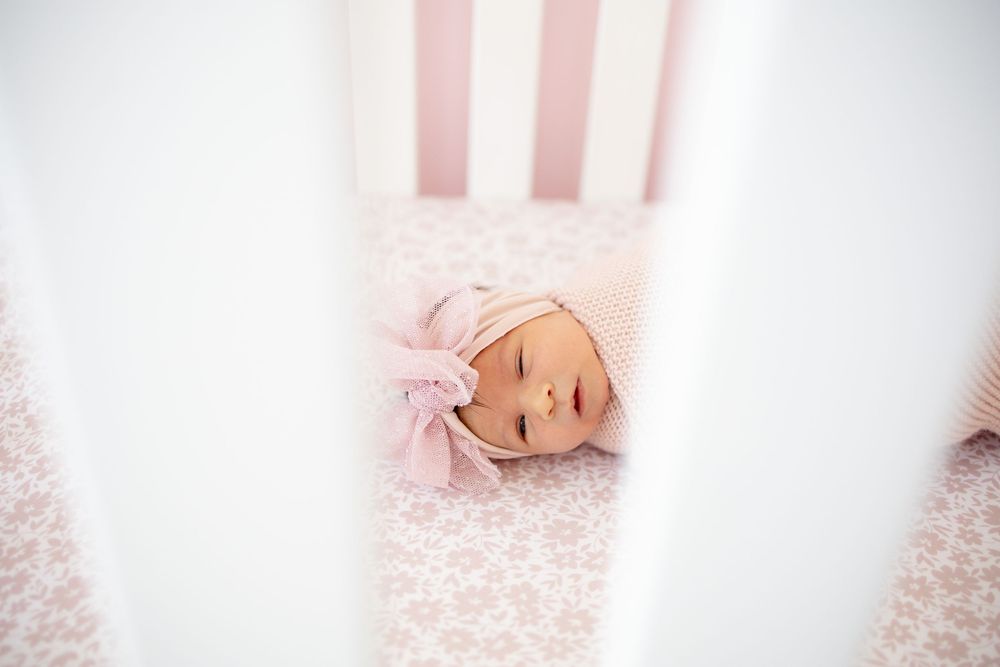 Newborn baby girl with pink bow headband sleeping peacefully on floral crib sheet inside white wooden crib.