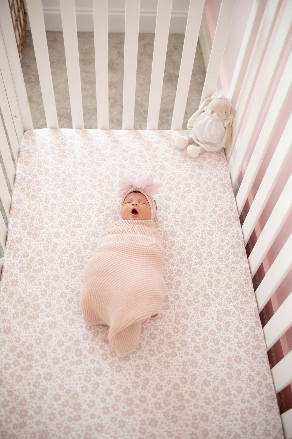 Newborn baby girl in pink swaddle with bow headband yawning in white crib with floral sheet and bunny toy.