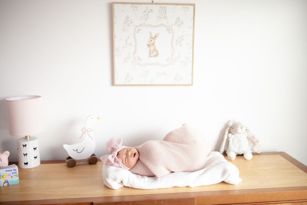Newborn baby girl in pink swaddle sleeping on dresser surrounded by bunny nursery decor and soft toys.