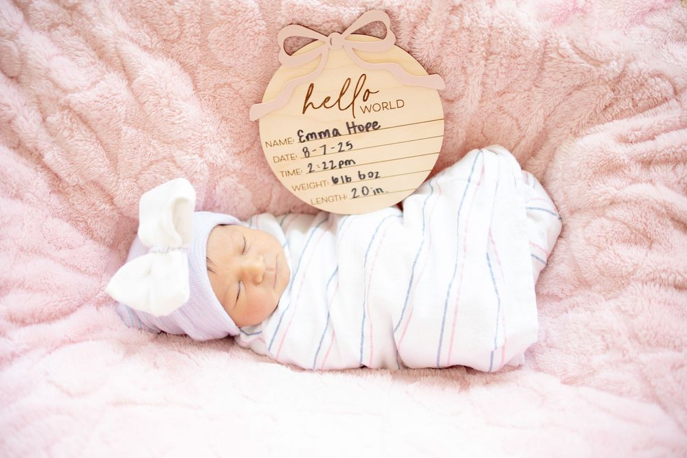 Sleeping newborn baby girl swaddled in white stripes with hello world wooden birth stats sign on pink blanket.