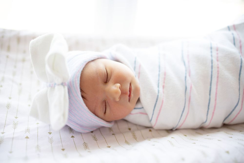 Peaceful newborn baby sleeping swaddled in white striped hospital blanket with white knotted bow hat.