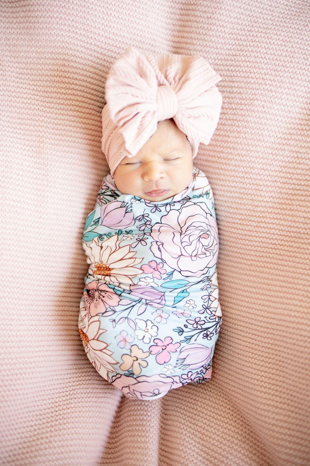 Newborn baby girl swaddled in floral blanket wearing pink bow turban headband sleeping on pink knit blanket.
