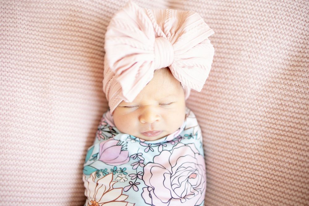 Close-up of sleeping newborn baby girl in floral swaddle and pink oversized bow turban headband.