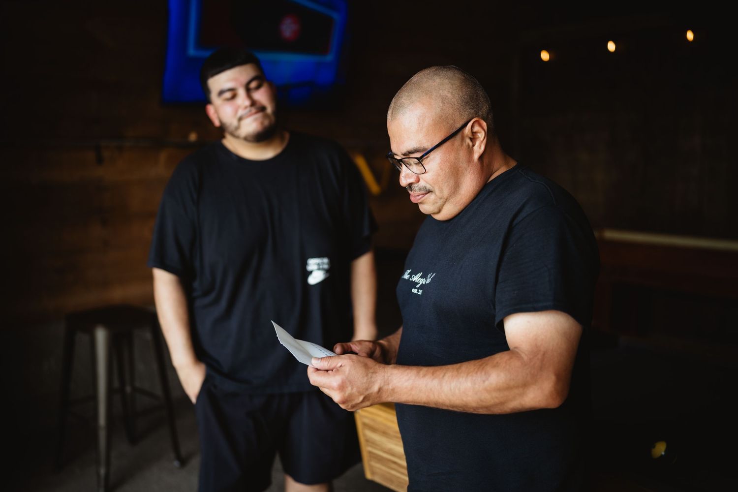 Two people in black t-shirts look at paperwork together in a dimly lit space.