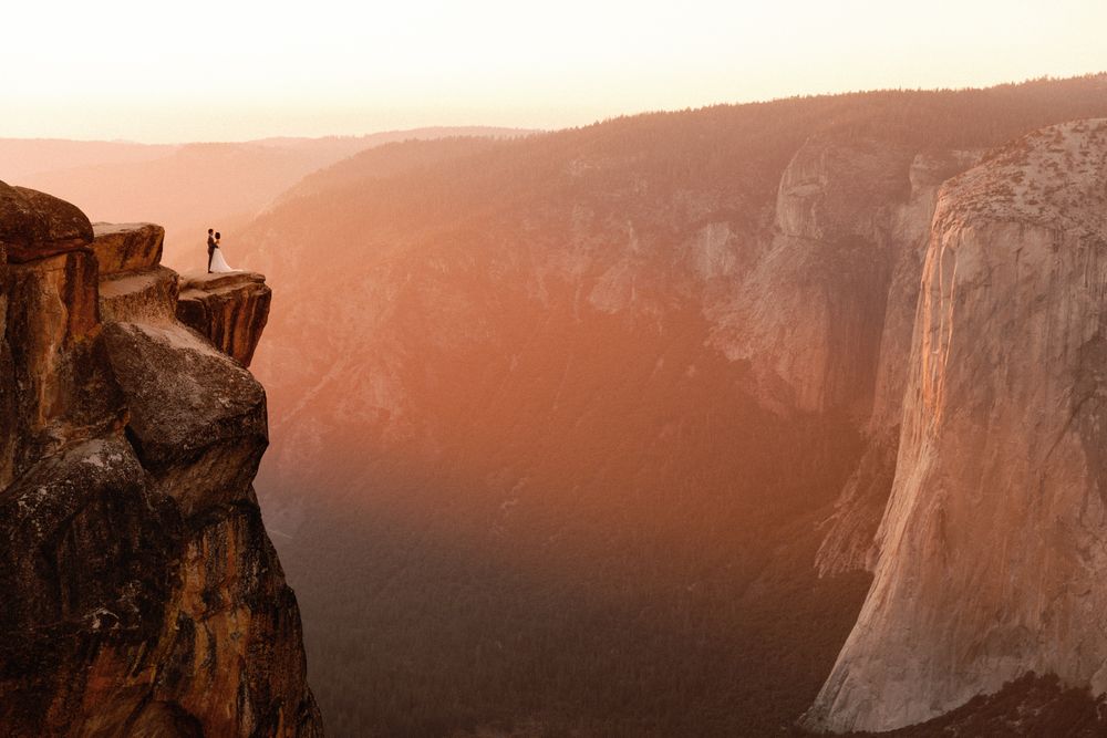 Tiny figure standing on a dramatic cliff edge at sunset overlooking Yosemite Valley with El Capitan in view.