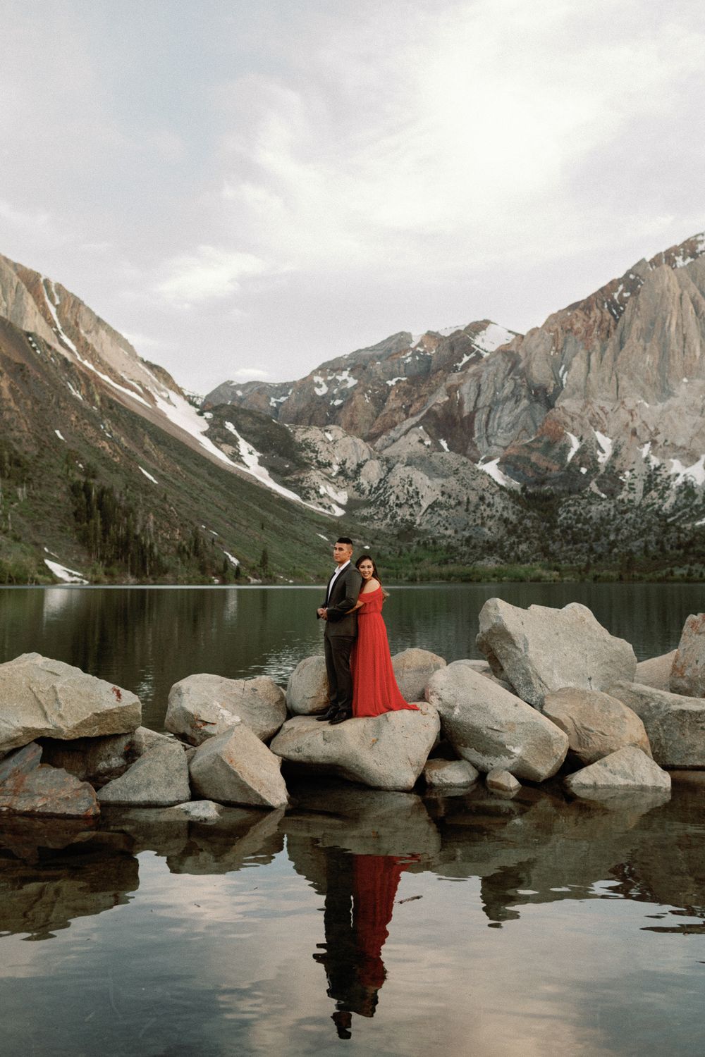 Couple in formal wear posing on rocky shore with snow-capped mountains reflected in lake.